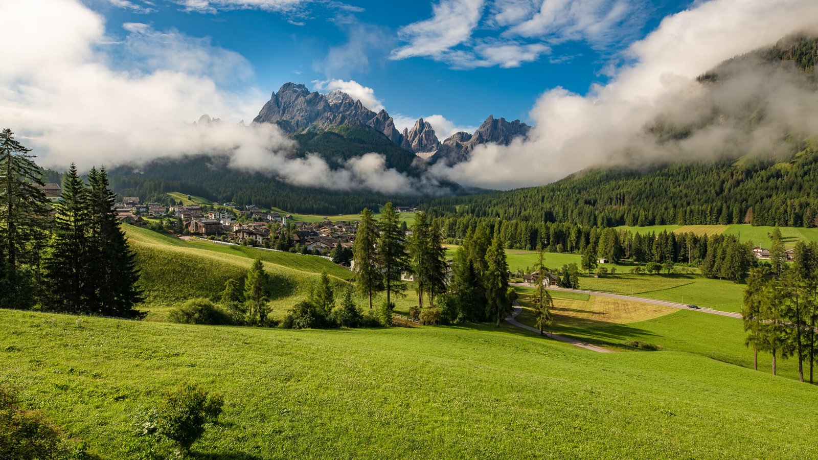 Grüne Wiesen und Nadelwald vor den Berggipfeln der Dolomiten bei Sexten, mit Wolken über der Landschaft.